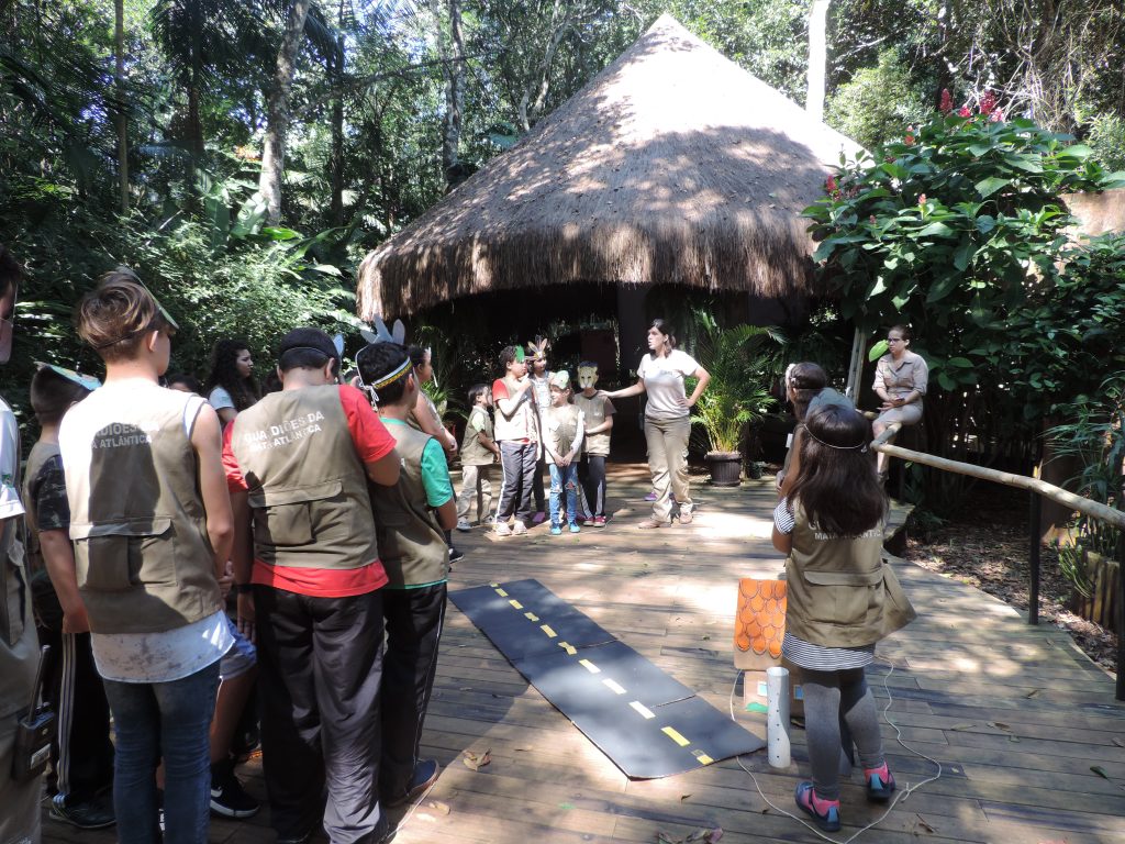 Um grupo de crianças e adolescentes, todos usando coletes de exploradores, participa de uma atividade ao ar livre em um ambiente cercado por vegetação densa.