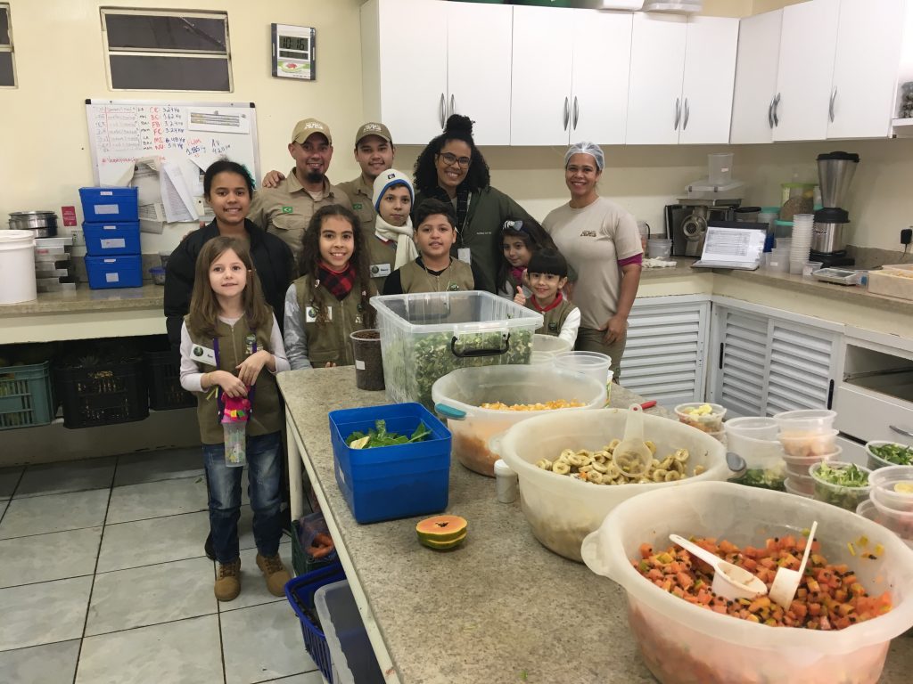 Um grupo de crianças e adultos, todos sorrindo, posam para uma foto em uma cozinha industrial. As crianças estão vestindo uniformes de exploradores, indicando que fazem parte de um programa educativo. 