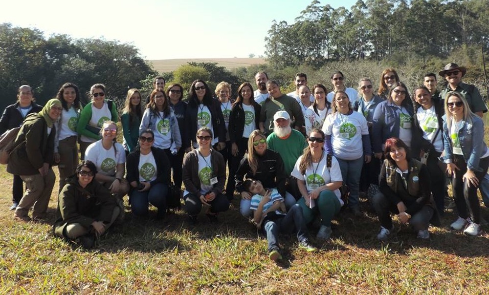 Grupo de pessoas do Projeto SOS Fauna está reunido para uma foto em um ambiente natural, durante uma atividade de campo relacionada à conservação ambiental. O ambiente ao redor é um campo aberto com vegetação e árvores ao fundo, sugerindo um dia ensolarado e propício para atividades ao ar livre.