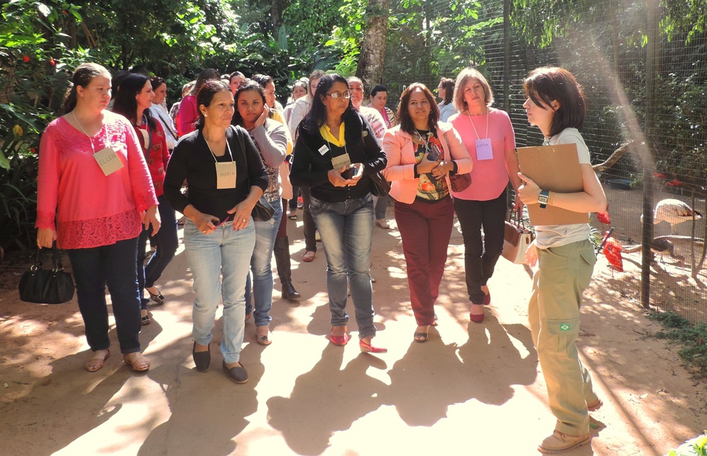 Um grupo participando de uma visita guiada no Parque das Aves. A guia, que está à direita da imagem, segura uma prancheta e está explicando algo para os visitantes. O grupo está atento e interessado, e a vegetação ao redor é ambiente natural e agradável.