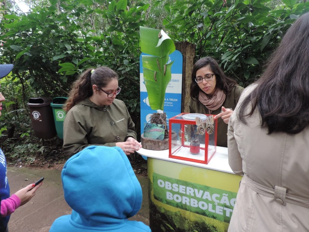 Duas mulheres em uma estação educativa ao ar livre, mostrando insetos dentro de uma caixa de vidro vermelha para um grupo de visitantes. A estação é dedicada à observação de borboletas.