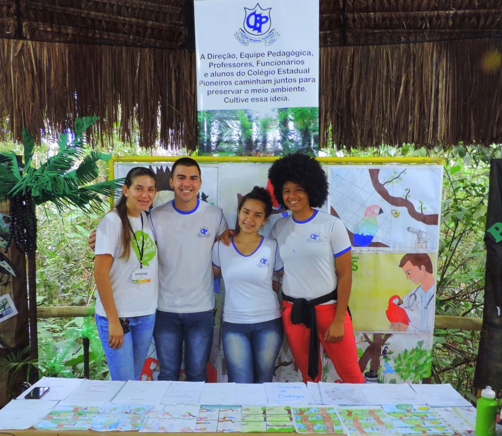 Quatro estudantes do Colégio Estadual Pioneiros posando juntos em uma exposição sobre preservação ambiental. Ao fundo, há cartazes com desenhos e mensagens sobre conservação.