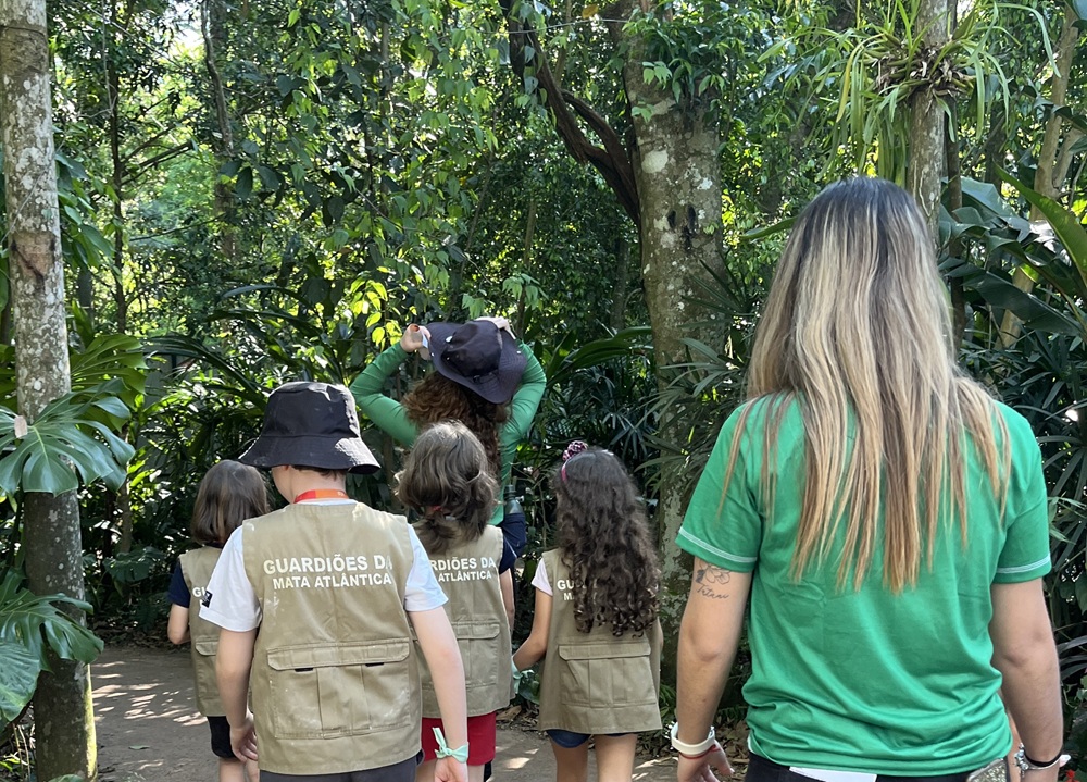 Crianças usam coletes escritos "Guardiões da Mata Atlântica" caminham por uma trilha no Parque das Aves, acompanhados por educadores.