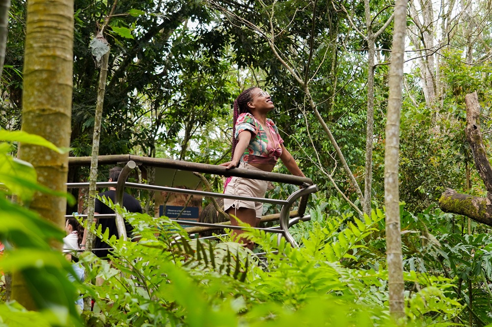 Mulher sorrindo de olhos fechados em uma plataforma de observação no Viveiro Cecropia, cercada por vegetação exuberante da Mata Atlântica.