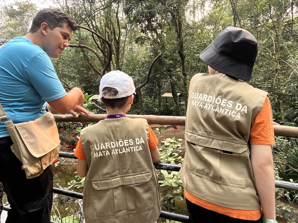 Um guia com camiseta azul e bolsa bege conversa com duas crianças usam coletes com a inscrição "GUARDIÕES DA MATA ATLÂNTICA" nas costas. Eles estão em um mirante de madeira, observando a natureza ao redor de um ambiente de floresta.