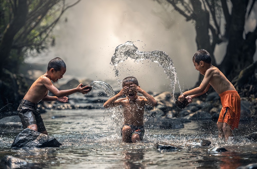 Três meninos brincando e se molhando em um riacho, cercado por pedras e árvores, com água sendo tocada no ar.