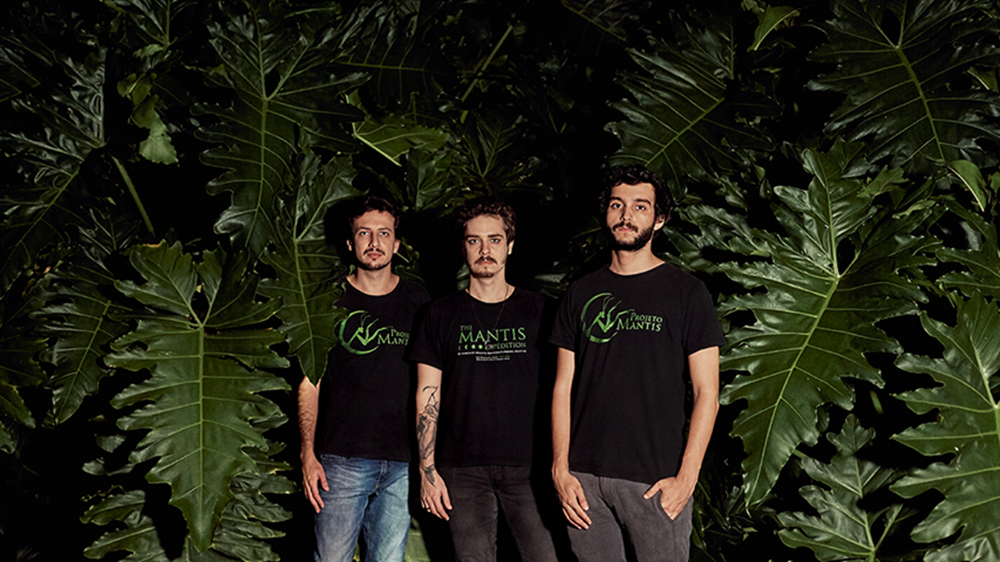 Três homens posando lado a lado, usando camisetas pretas com o logotipo do Instituto Mantis, em frente a um fundo com grandes folhas verdes tropicais.