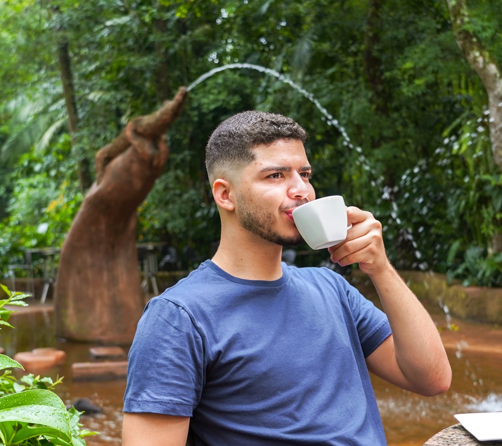 Homem com camisa azul tomando café em um ambiente ao ar livre, com uma fonte em formato de jacaré e vegetação ao fundo.