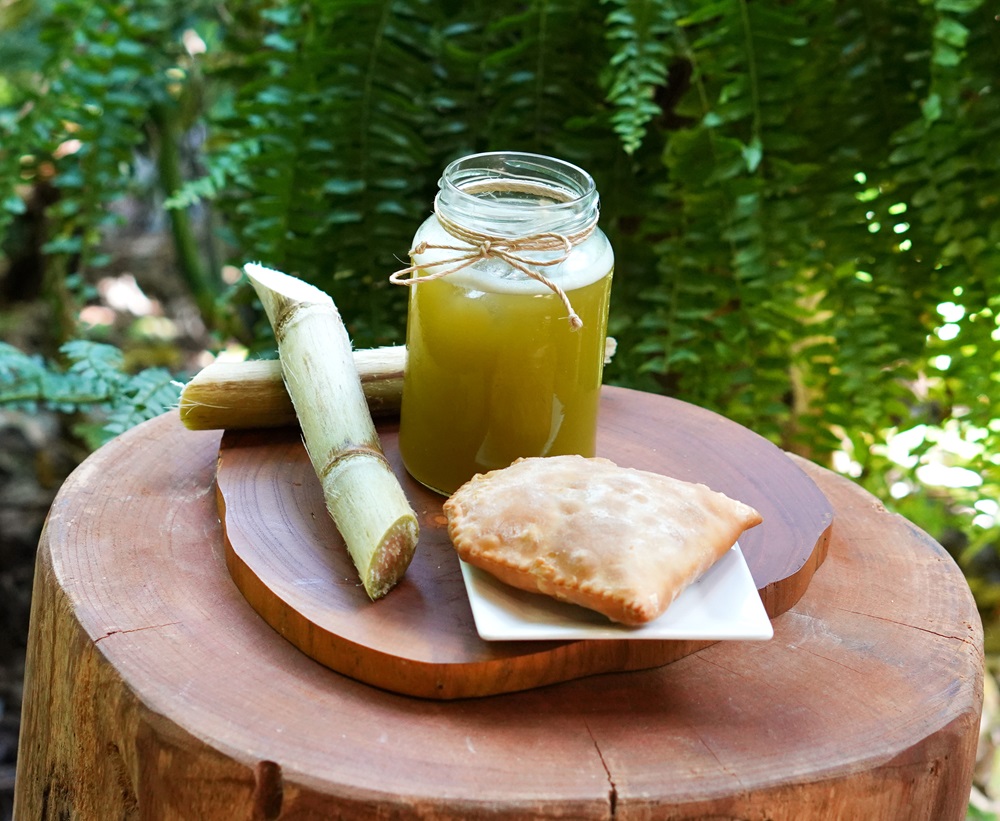 Pastel dourado e crocante ao lado de um copo de caldo de cana em um pote de vidro decorado com barbante, sobre uma tábua de madeira com vegetação ao fundo.