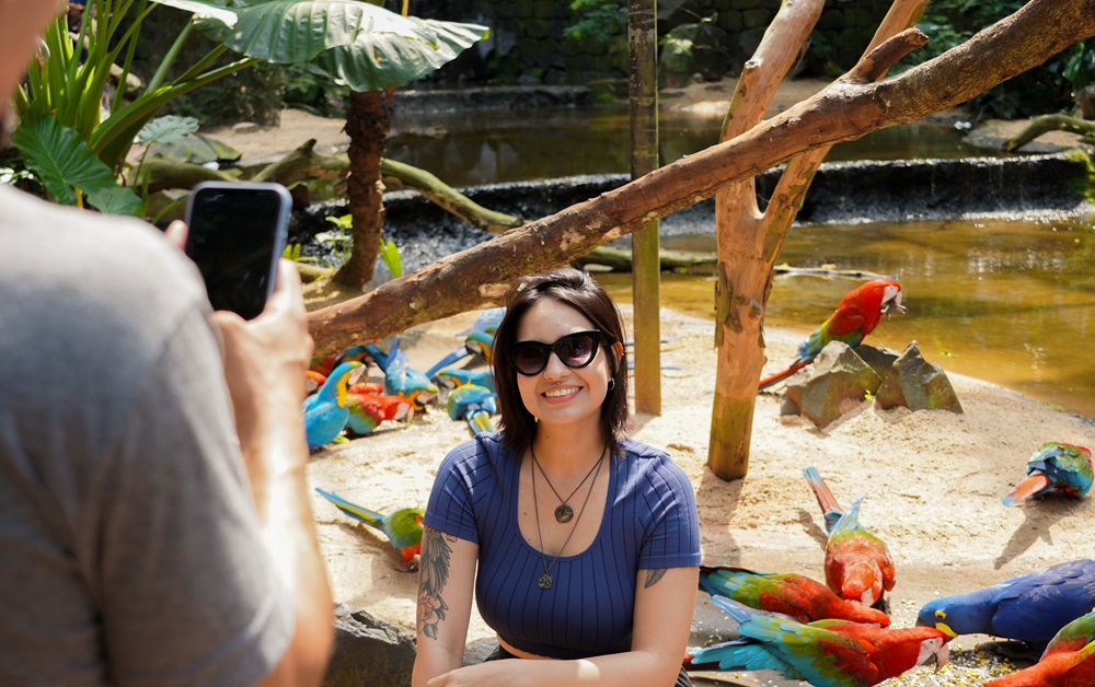 Mulher sorrindo em frente às araras no viveiro, com uma cena colorida de pássaros ao fundo. No Parque das Aves, em Foz do Iguaçu