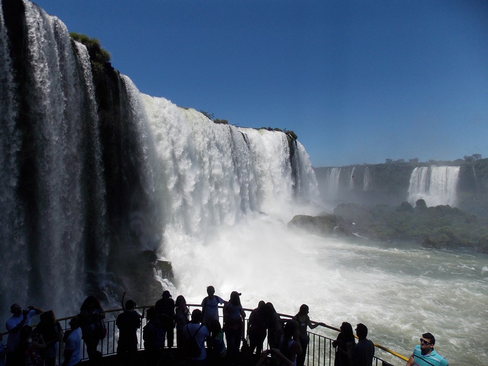 Turistas observando as Cataratas do Iguaçu de uma passarela, cercados por um céu azul e o som das quedas d'água, em Foz do Iguaçu