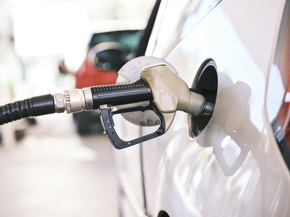 Bico de bomba de combustível abastecendo um veículo branco em um posto de gasolina, com outro carro desfocado ao fundo.