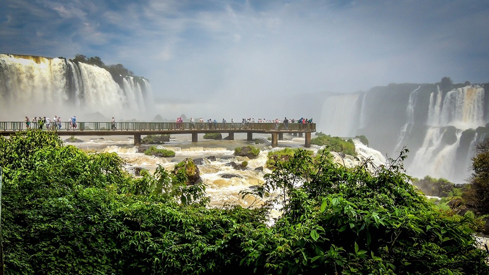 Vista panorâmica das Cataratas do Iguaçu com destaque para a passarela de observação, cercada por vegetação exuberante e quedas d'água impressionantes ao fundo.