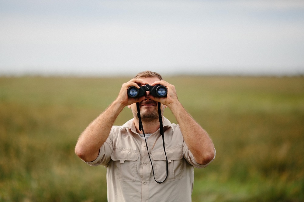 Um homem em uma paisagem de campo aberto utilizando binóculos para observar aves à distância, com expressão especializada. Ele veste uma camisa bege e tem um fundo de vegetação verde desfocado.