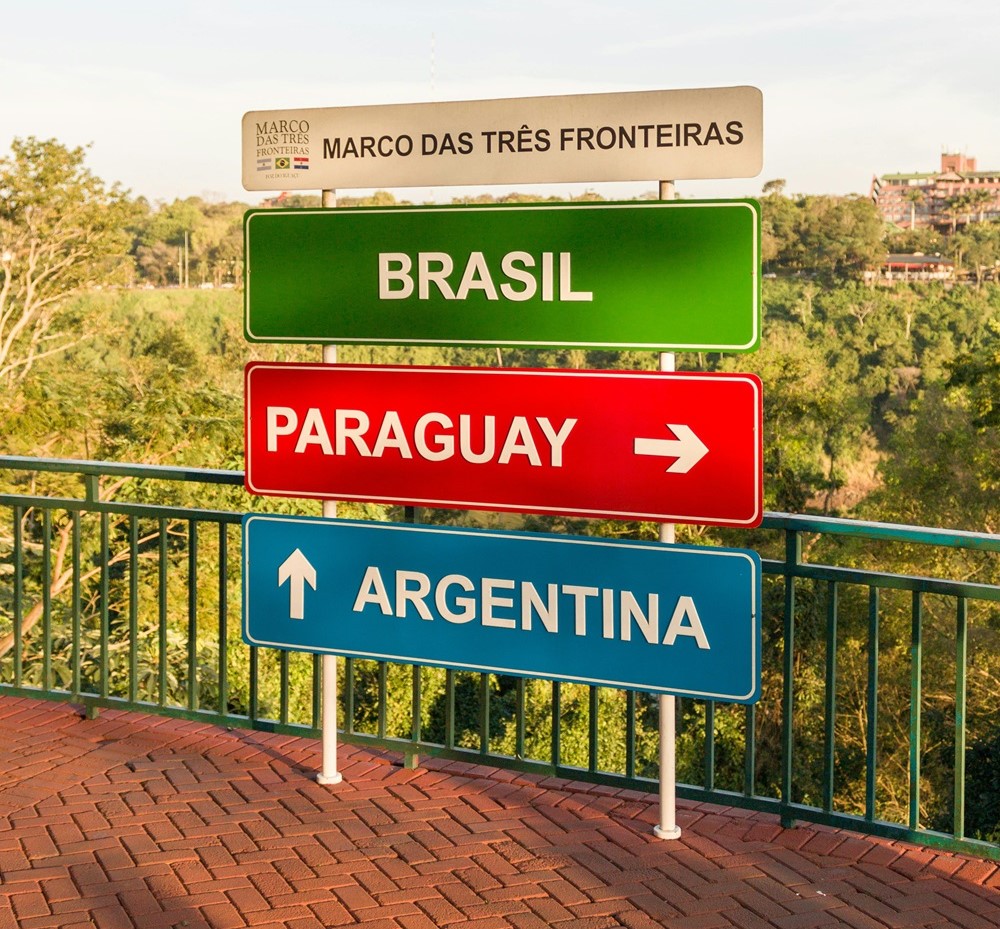 Placa indicativa no Marco das Três Fronteiras, em Foz do Iguaçu, com três sinais coloridos representando Brasil (verde), Paraguai (vermelho, com seta para a direita) e Argentina (azul, com seta para cima). Ao fundo, vegetação e céu claro compõem a paisagem.