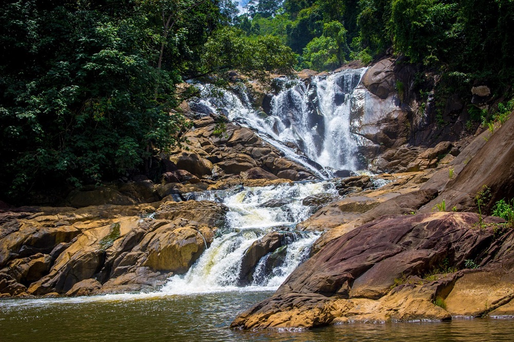 Vista de uma cachoeira em meio à natureza com rochas e vegetação ao redor. A água flui em cascatas formando pequenos poços naturais.