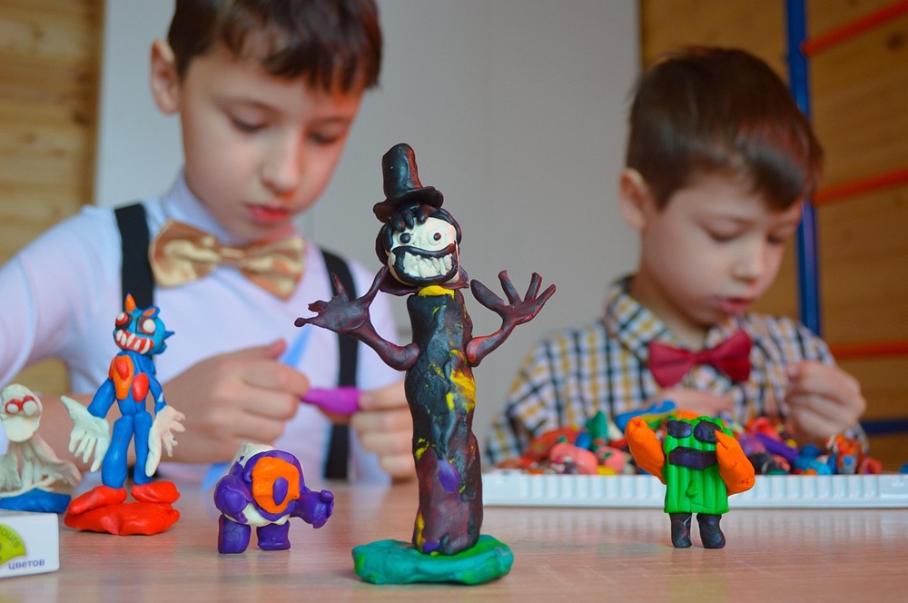 Duas crianças equipamentos enquanto modelam esculturas coloridas de massinha em uma mesa, com diversas figuras criativas no primeiro plano.