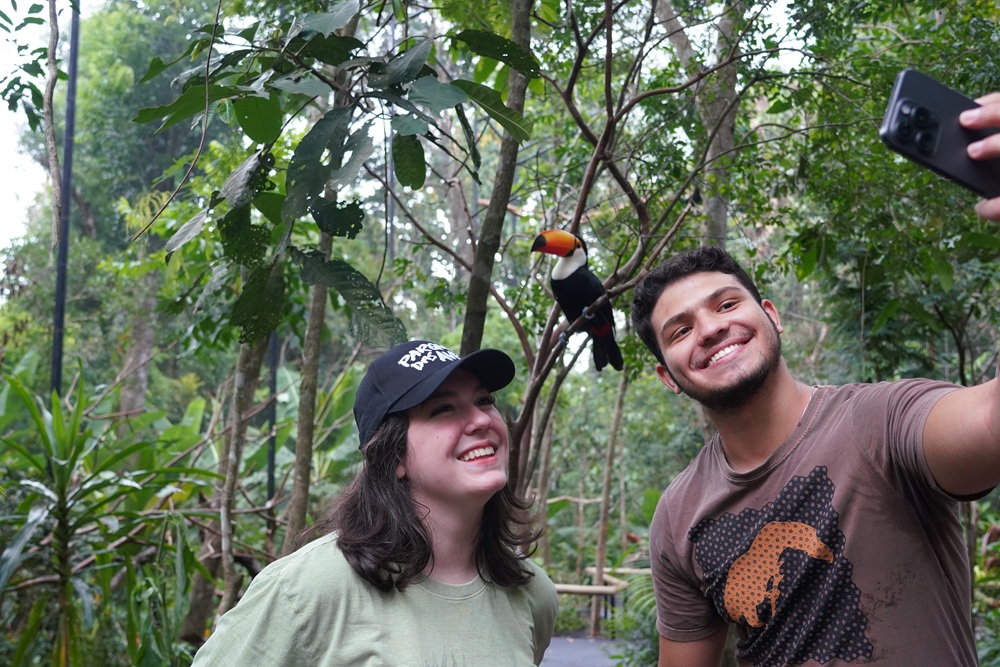 Dois visitantes sorridentes tiraram uma selfie em meio à vegetação do Parque das Aves. Ao fundo, um tucano pousado em um galho observa a cena.