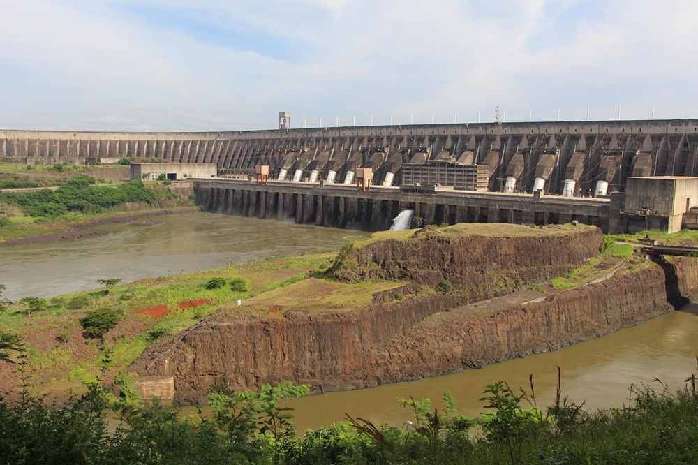 Vista panorâmica da Usina Hidrelétrica de Itaipu, com sua imponente estrutura de concreto e vertedouros liberando água. O cenário inclui o rio em primeiro plano, vegetação ao redor e o céu parcialmente nublado ao fundo.