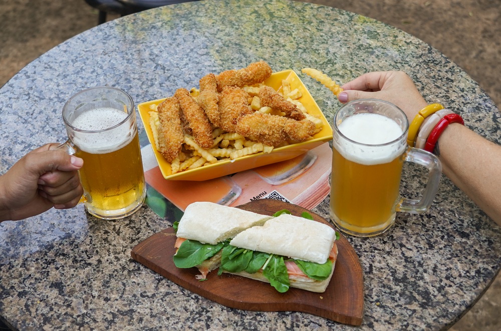 Mesa de granito com dois copos de chope, um sanduíche com salmão e rúcula, e uma porção de batatas fritas com iscas de frango empanadas. Duas pessoas brindam enquanto apreciam uma refeição ao ar livre.