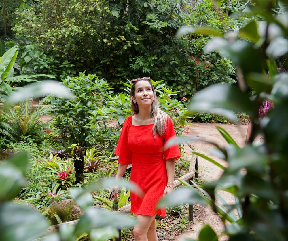 Mulher de vestido vermelho caminha por uma trilha cercada pela vegetação da Mata Atlântica no Viveiro Borboletas e Beija-flores, com um olhar sereno enquanto aprecia o ambiente ao redor.