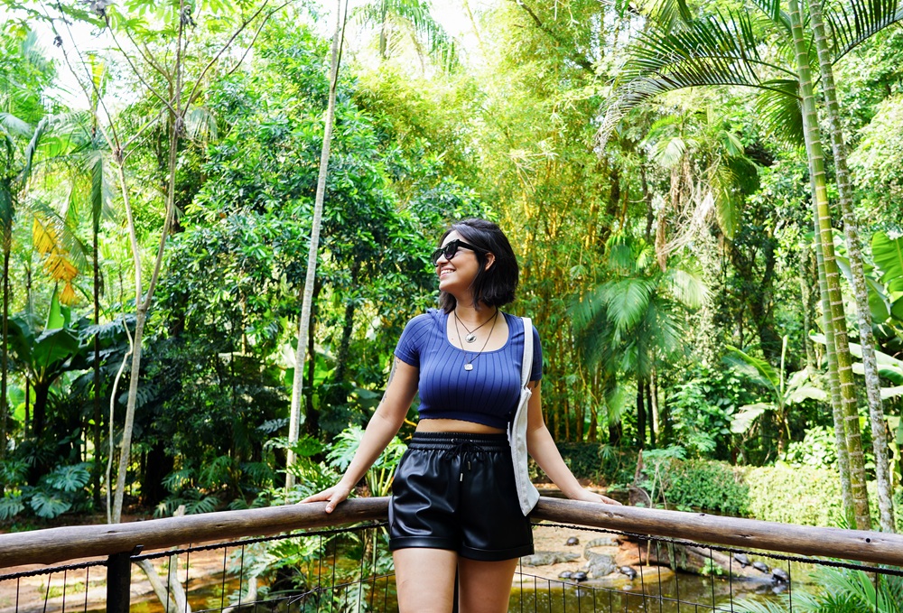 Mulher sorridente, usa blusa azul e shorts pretos, encostada em um corredor de madeira no Viveiro dos Crocodilos, cercada por um vegetação densa da Mata Atlântica e iluminada pelo sol.