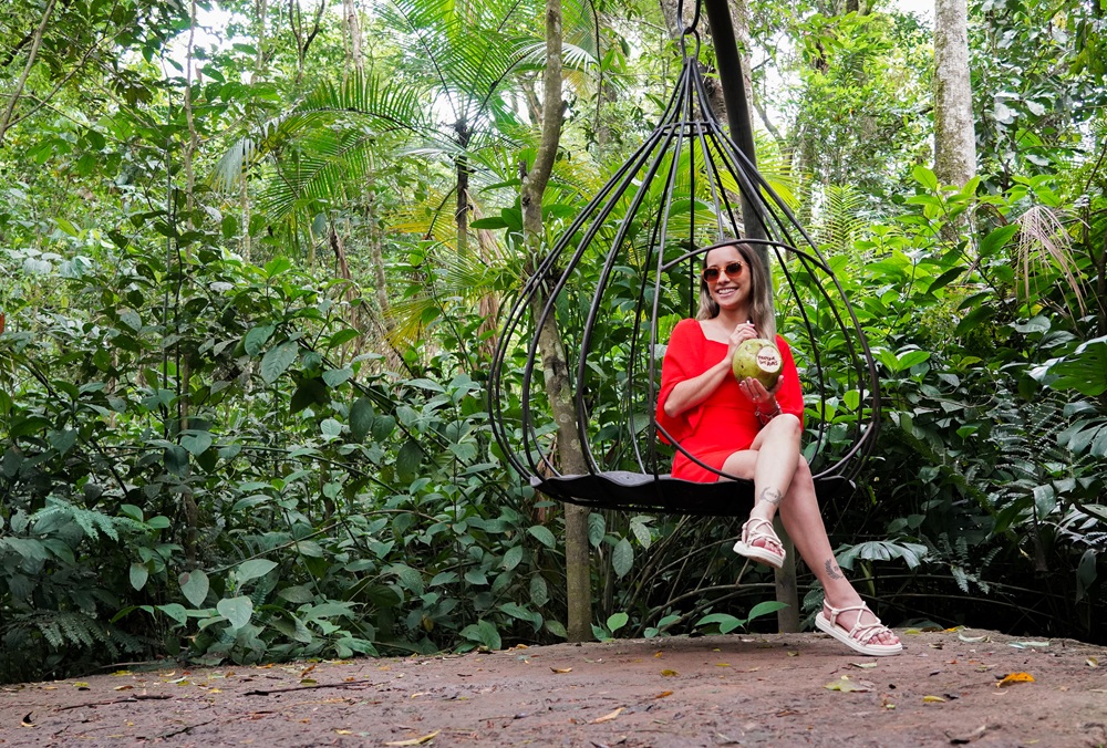 Mulher de vestido vermelho e óculos de sol relaxa em um balanço suspenso no meio da Mata Atlântica, segurando um coco.