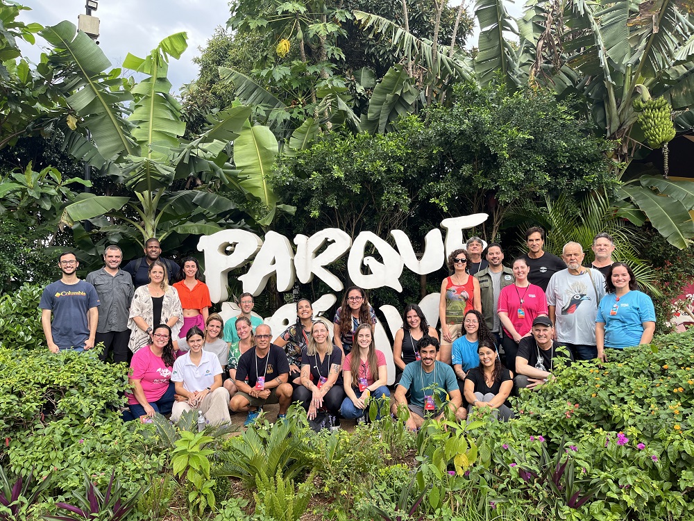Um grupo de pessoas está posando em frente a um grande letreiro branco que diz "Parque das Aves". Elas estão cercadas por uma vegetação exuberante com muitas plantas verdes e grandes folhas. Todos parecem felizes e estão sorrindo para a câmera.