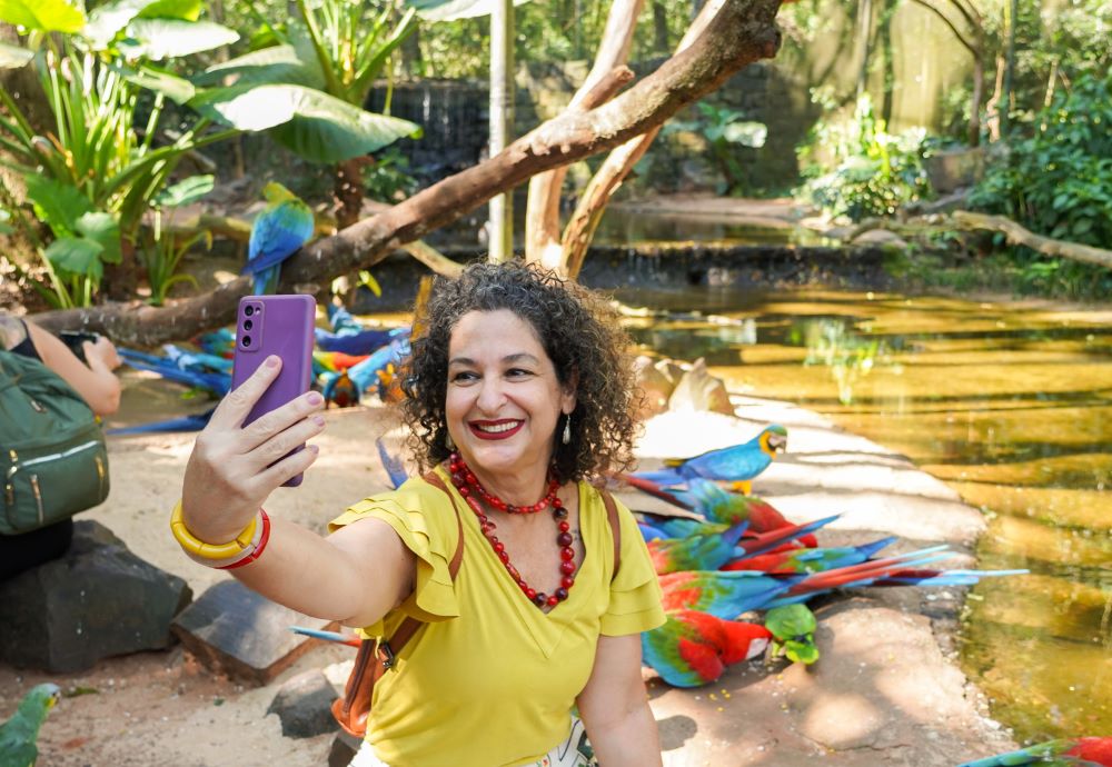 Uma mulher sorridente, vestindo uma blusa amarela e colares vermelhos, está tirando uma selfie com um celular roxo em um ambiente de floresta tropical. Ao fundo, várias araras coloridas estão pousadas em troncos e no chão, com vegetação densa ao redor.