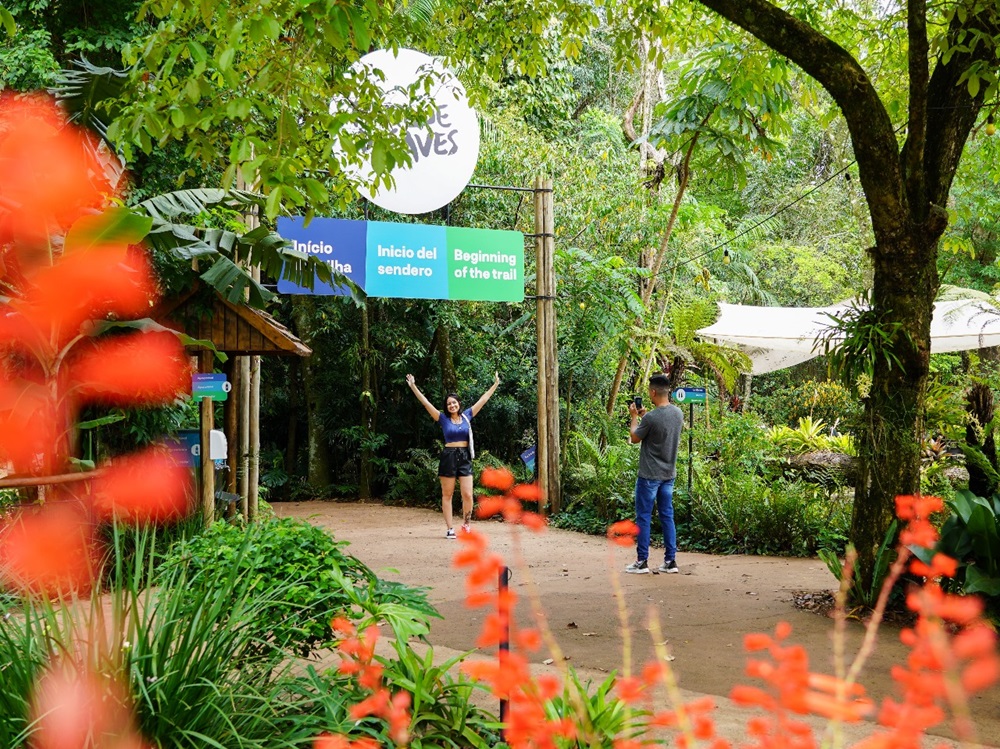 pessoa posando de braços abertos sob uma placa de início da trilha no Parque das Aves, enquanto outra pessoa tira uma foto. A área é cercada por vegetação tropical densa, com flores vermelhas em primeiro plano.