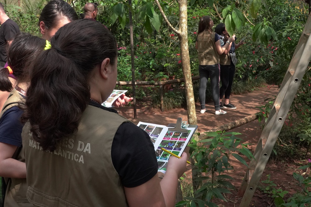 Crianças com coletes "Guardiões da Mata Atlântica" observando uma folha de papel, participando de uma atividade ao ar livre.