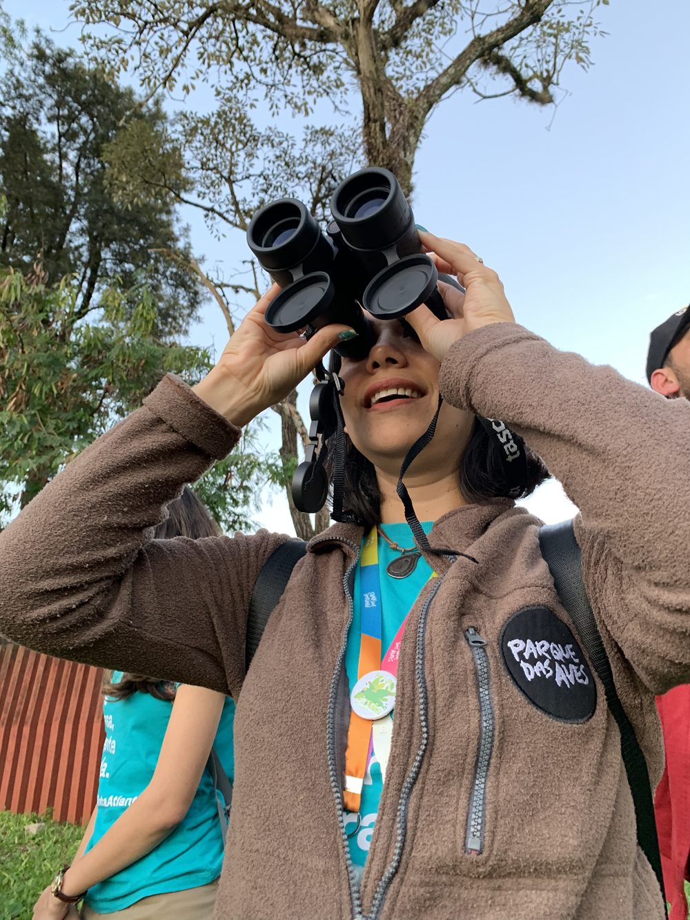 Mulher usando um binóculo para observar o céu, vestindo um casaco com o logo do Parque das Aves, ao ar livre, com árvores ao fundo.