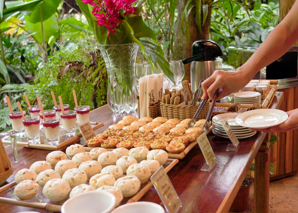 Uma mesa de coffee break em um ambiente natural com uma variedade de pães de queijo, lanches e sobremesas, enquanto uma pessoa serve-se utilizando um pegador e segura um prato. Flores decorativas estão ao fundo, adicionando um toque de cor ao ambiente.