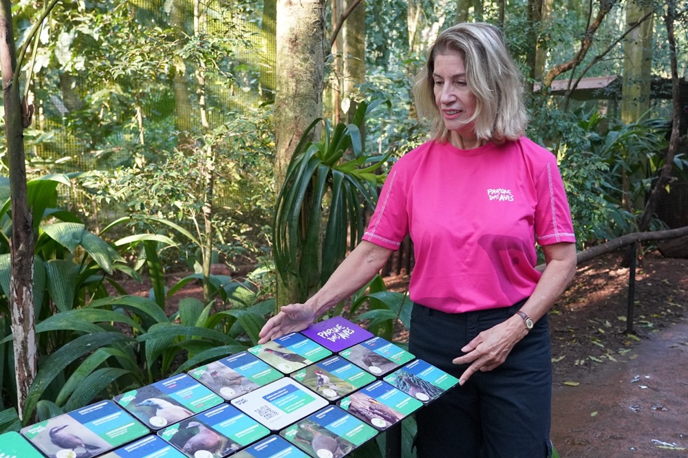 Uma mulher vestindo uma camiseta rosa do Parque das Aves está em pé em um ambiente de floresta tropical. Ela está apontando para uma mesa com várias placas informativas sobre aves. A vegetação ao redor é densa, criando um ambiente natural e educativo.