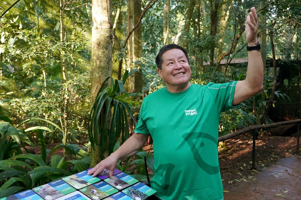 Um homem sorridente, vestindo uma camiseta verde do Parque das Aves, está em pé em um ambiente de floresta tropical. Ele está apontando para cima com uma das mãos enquanto explica algo. Na mesa à sua frente, há várias placas informativas com imagens de aves.