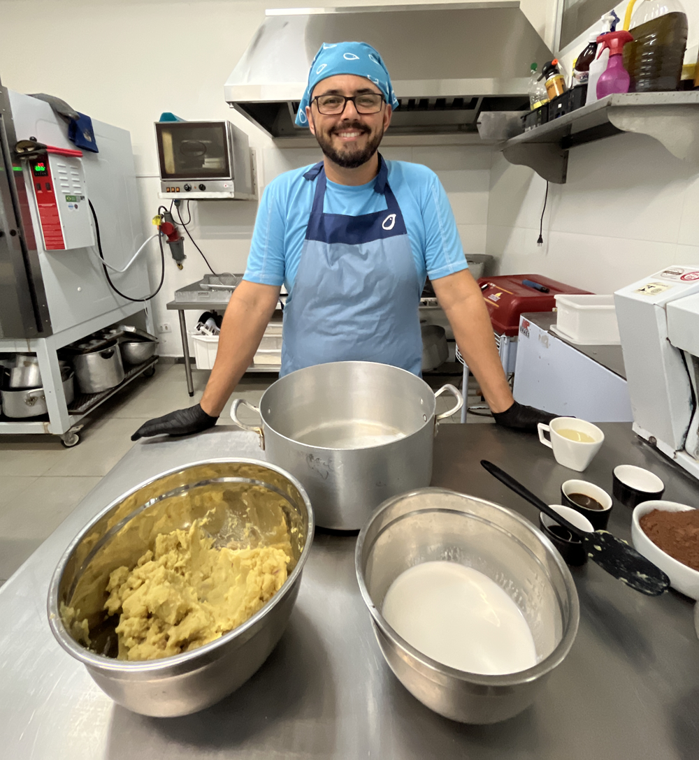  Elisier Taschetto, sorridente, usando avental azul e touca, em uma cozinha, com ingredientes do brigadeiro vegano à sua frente.