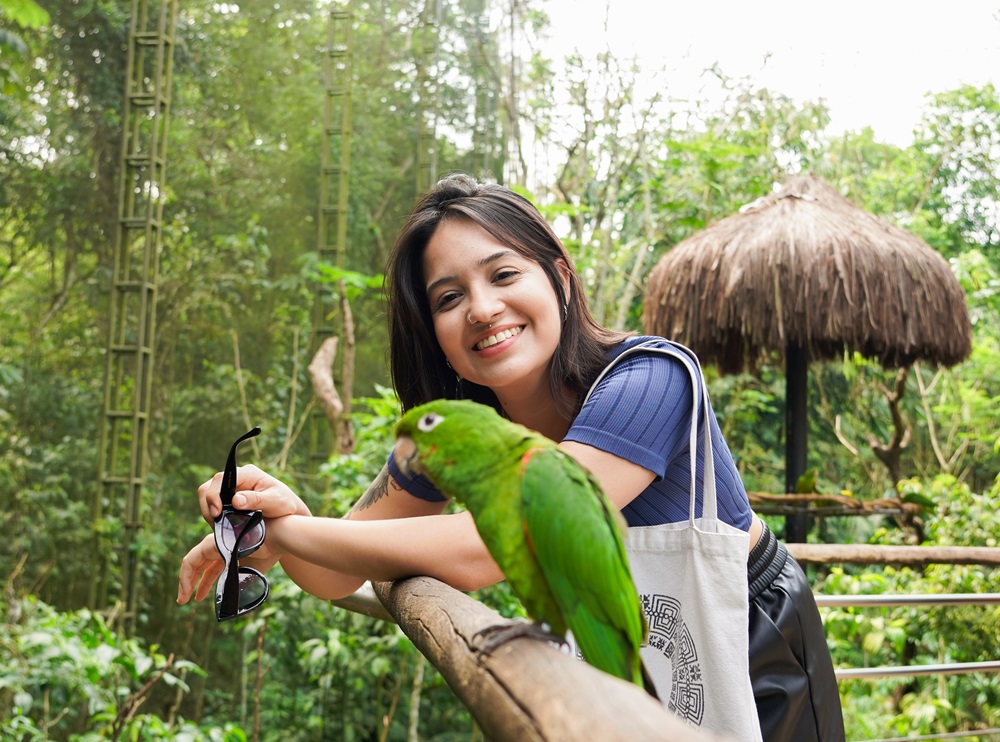 Visitante sorrindo enquanto observa de perto um periquito pousado em uma cerca de madeira no viveiro Cecropia do Parque das Aves.