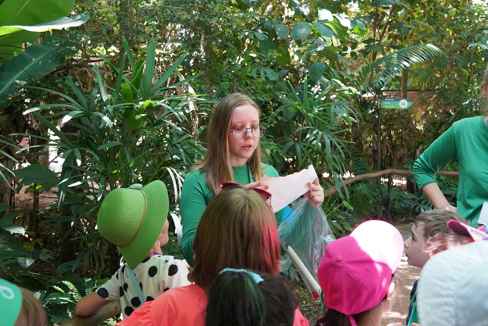 Educadora ambiental, durante a colônia de férias, explicando uma atividade para um grupo de crianças reunidas em meio à vegetação densa do Parque das Aves.