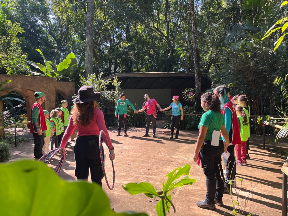 Crianças e educadores durante a colônia de férias, organizados em um círculo ao ar livre, participando de uma dinâmica educativa em uma área arborizada do Parque das Aves.