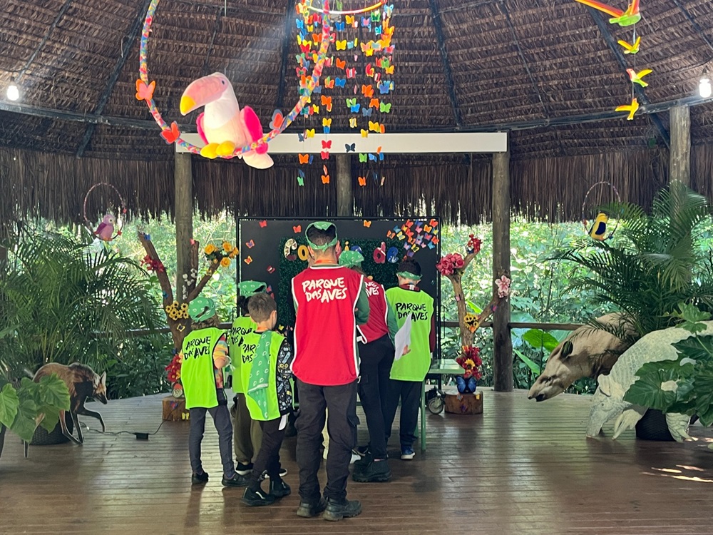 Crianças e educadores reunidos durante a colônia de férias, sob uma estrutura coberta com teto de palha, decorados com elementos coloridos e educativos, participando de uma atividade lúdica no Parque das Aves.