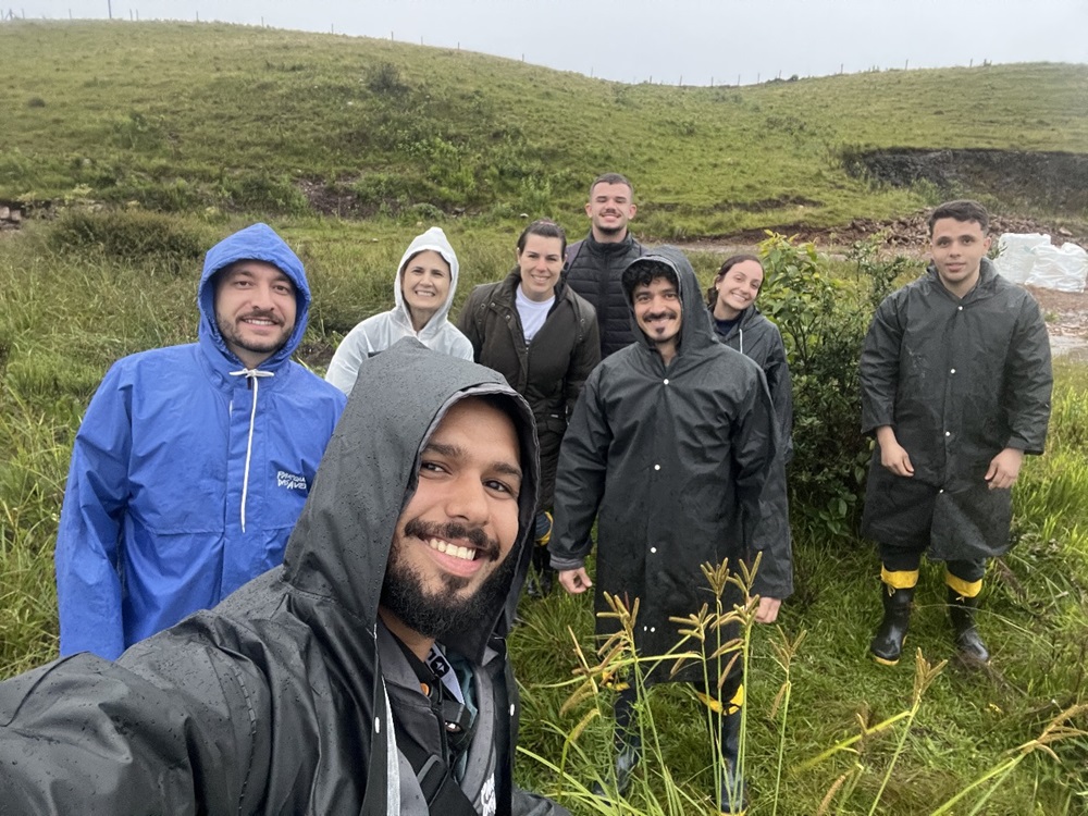 Grupo de oito pessoas usando capas de chuva em uma área de campo aberto, posando sorridentes para uma selfie em meio à vegetação úmida, em busca da perereca-rústica