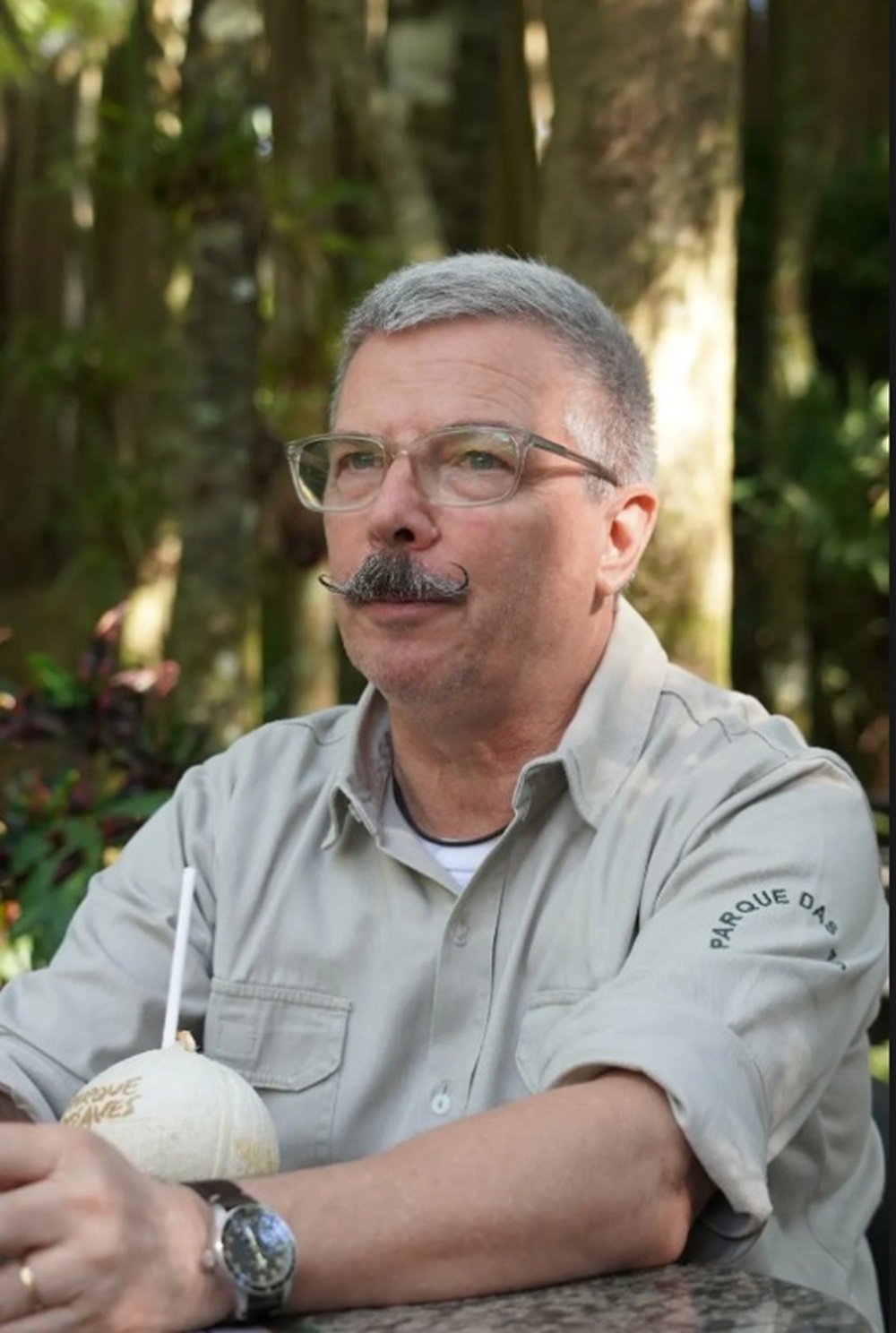 Homem de cabelo grisalho e bigodeado estilizado, usa uniforme bege do Parque das Aves, sentado em uma mesa ao ar livre com um coco personalizado com o logotipo do Parque.