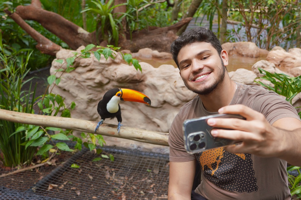 Jovem turista sorridente tirando uma selfie ao lado de um tucano sobre um galho, com vegetação exuberante ao fundo no Parque das Aves. no viveiro dos tucanos.