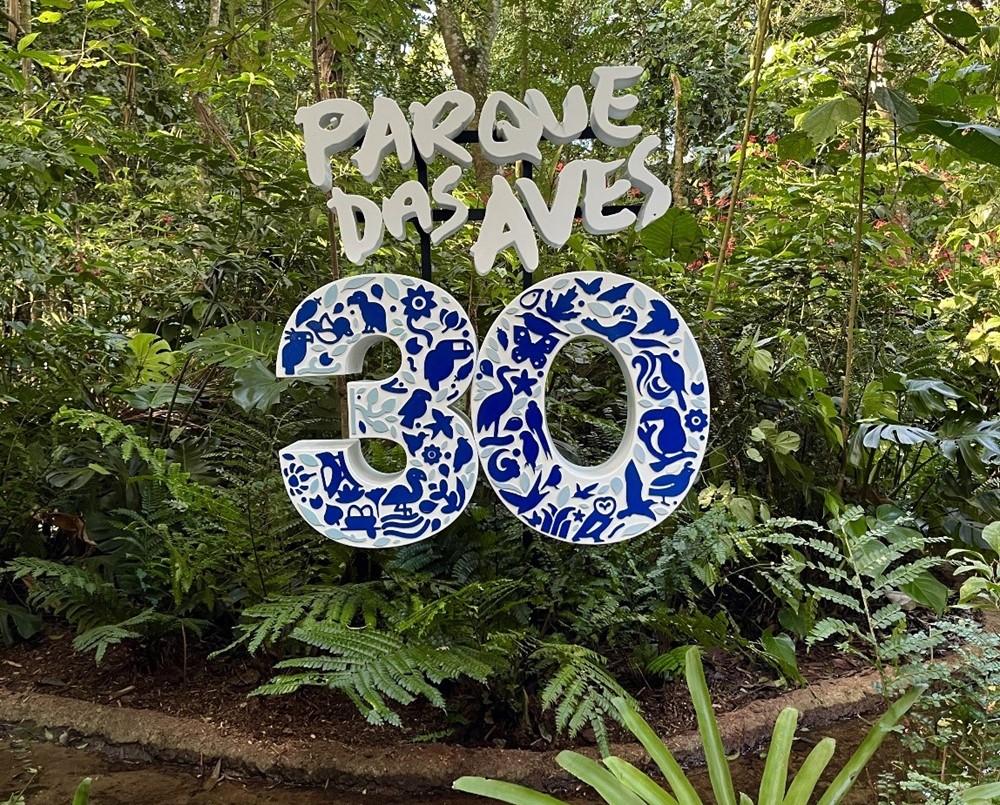 Estrutura comemorativa com o texto 'Parque das Aves 30', decorada com desenhos azuis de aves e outros elementos, posicionada em meio à vegetação do Parque