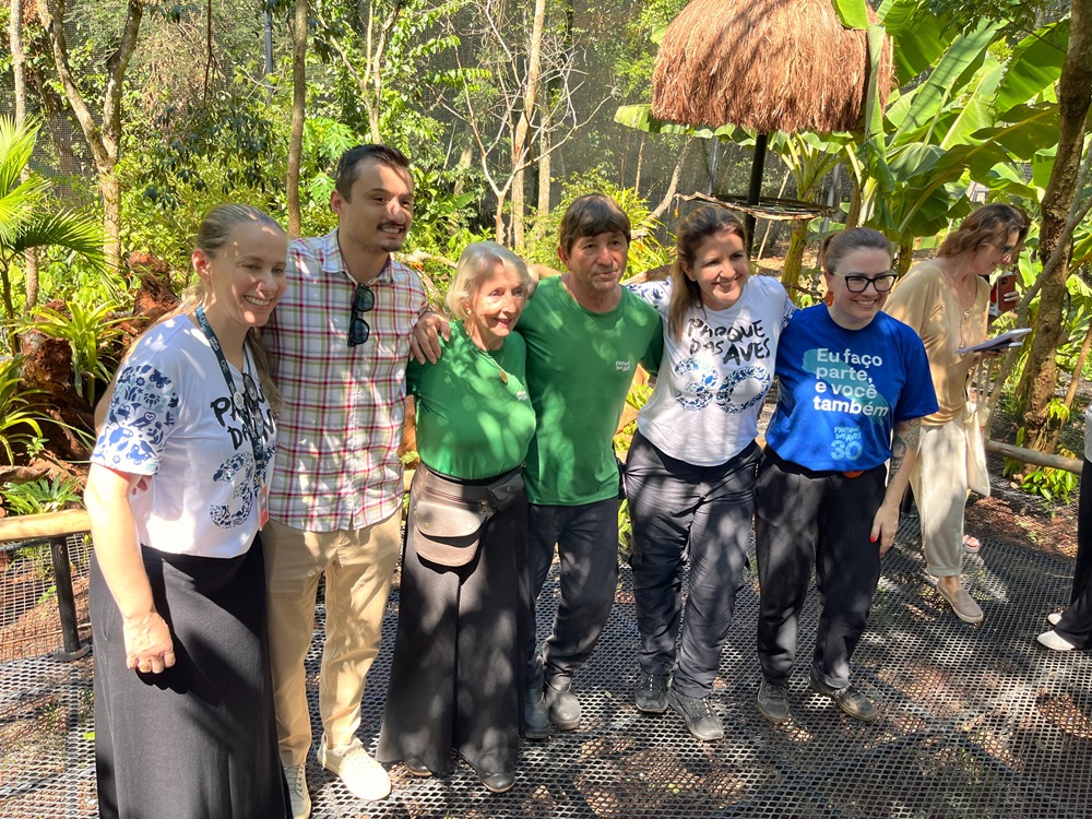  Grupo de seis pessoas posando para uma foto no Parque das Aves, no viveiro dos tucanos, cercadas por vegetação nativa e estruturas rústicas de madeira e palha. Duas mulheres usam camisetas comemorativas de 30 anos do Parque das Aves.